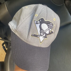 Pittsburgh Penguins keps från Reebok - Snygg Pittsburgh Penguins keps från Reebok med broderad logga framtill och text bak. Grått tyg med svart skärm och detaljer i vitt och gult. Perfekt för dig som älskar hockey och vill visa vilket lag du hejar på.