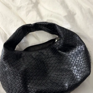 Svart handväska skinnimitation - Säljer en svart handväska med rundad form och flätad struktur. Väskan har dragkedja upptill och är gjord i skinnimitation