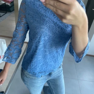 Blå spetsblus från Flash Woman - Supersöt blå blus i spets från Flash Woman. Blusen har trekvartsärmar och är helfodrad i kroppen men transparent på ärmarna. Blommigt spetsmönster och rak passform, perfekt till jeans eller kjol för en trendig look.