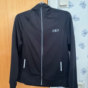 Svart träningskofta från OTP med luva i storlek L - Svart träningskofta från OTP med hel dragkedja och luva, storlek L. Jackan har reflekterande detaljer på fickor och axel, samt logga på bröstet. Perfekt för utomhusaktiviteter och träning. Köparen står alltid för frakten