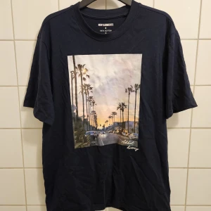 Mörkblå t-shirt med LA-print New Garments - Mörkblå t-shirt från New Garments i 100% bomull. Framsidan har ett stort tryck med palmer och solnedgång från Abbot Kinney i Los Angeles. Relaxed passform och rund hals, perfekt för en chill streetstyle look. Tröjan köptes för 200 kr. Pris går att diskutera och skriv gärna frågor om det finns. Den har knappt använts och är i väldigt bra skick.