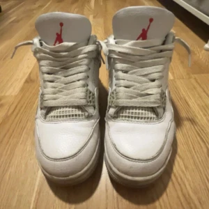 Nike Air Jordan 4 White Cement - Säljer ett par Nike Air Jordan 4 White Cement med klassisk vit ovandel i skinn, meshpaneler och grå detaljer med stänk på sulan. Ikonisk Jumpman-logga i rött och silver på plösen och hälen. Perfekta för dig som vill sticka ut med en tidlös sneaker.