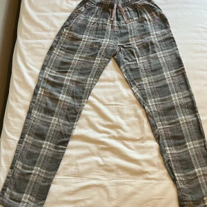 Grå rutiga mjukisbyxor med snörning - Mysiga grå mjukisbyxor med vitt, rosa och grått rutmönster. Byxorna har elastisk midja med snörning och raka ben. Perfekta för chill dagar hemma eller när du vill vara extra bekväm. Tillverkade i mjukt bomullstyg. Har två par ett i storleken 158+164 och ett par i 170 
