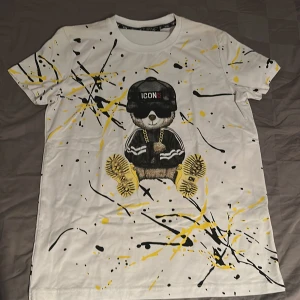 Vit  t-shirt med björntryck - Vit t-shirt från  med coolt tryck av en björn i streetwear, keps och guldkedja. T-shirten har korta ärmar och är täckt av svarta och gula färgstänk för en unik look. Perfekt för dig som vill sticka ut med en trendig och lekfull stil.