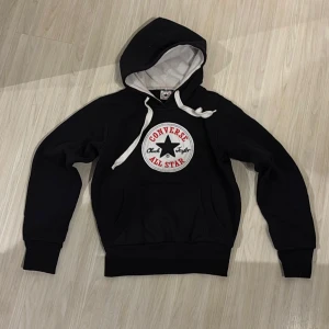 Svart Converse hoodie med logga - Svart hoodie från Converse med stor broderad All Star-logga på bröstet. Tröjan har vit snörning i huvan och en klassisk känguruficka framtill. Tillverkad i 100% bomull för en skön och avslappnad känsla.