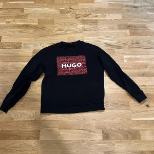 Svart sweatshirt från Hugo Boss med röd logga - Svart sweatshirt från Hugo med stor röd och svart logga framtill där det står HUGO i vitt. Tröjan har rund hals, långa ärmar och ribbade muddar. Materialet är mjuk bomull, perfekt för en chill och stilren look. Pris kan diskuteras vid snabb affär 