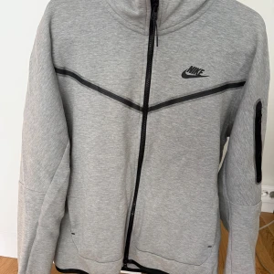 Grå zipjacka från Nike i storlek M - Snygg grå zipjacka från Nike med svarta detaljer och logga på bröstet. Jackan har hel dragkedja, hög krage och en ficka med dragkedja på ärmen. Perfekt för en sportig och avslappnad stil.