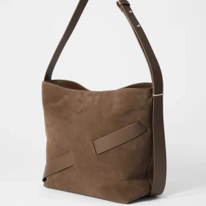 Nyskick! Brun väska i mocka med läderdetaljer - Bucketväska i mjuk mocka med kontrasterande läderdetaljer. Justerbart axelband. Guldfärgade detaljer och dold magnetstängning. Använd ett fåtal gånger. Nypris 1890kr för en månad sedan (säljs fortfarande)