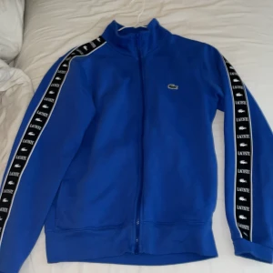 Blå Lacoste track jacket med logoband - Blå track jacket från Lacoste med dragkedja och klassisk ståkrage. Svarta och vita logoband med Lacoste-tryck längs ärmarna ger en sportig vibe. Jackan har lång ärm och är i mjukt bomullsmaterial. Perfekt för en chill och stilren look. Den är köpt för 1840kr på jd sports men nu finns den inte att köpa längre. Jag har använt den ett fåtal gånger ungefär 5-7 ggr men vill sälja den nu för behöver pengar 