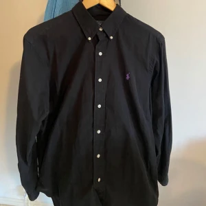 Svart skjorta från Ralph Lauren - Säljer nu min brors svarta skjorta från Ralph Lauren med klassisk button down-krage och lila broderad logga på bröstet. Skjortan har vita knappar hela vägen och är långärmad. Perfekt för dig som gillar stilrena plagg med en touch av färg. Säljs pga att han beställde för stor storlek och är då nämligen oanvänd (förutom att han testat den) Skjortan är perfekt nu till sommaren/hösten och passar bra på killar mellan 180-185 cm lång! vid frågor, tveka inte på att höra av er, alltid snabb respons!