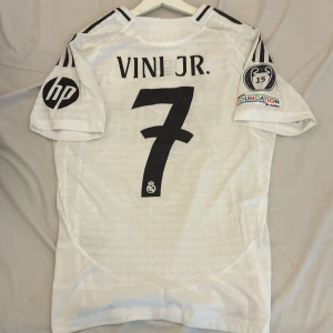 Real Madrid Vini Jr. 7 Champions League tröja - Säljer en Real Madrid fotbollströja från Adidas med Vini Jr. och nummer 7 från 2025 på ryggen. Tröjan är vit med svarta detaljer, Champions League-märke och HP-logga på ärmarna. Broderat klubbmärke och sponsortryck framtill. Tillverkad i lätt funktionsmaterial. Detta är tröjan spelarna använder (player version). Den säljs billigt pga att jag ite använder den längre. 