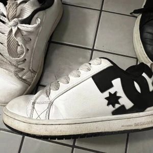 Vita DC sneakers med svart logga - Säljer ett par klassiska vita DC sneakers med stor svart DC-logga på sidan. Skorna har breda vita snören, svart innerfoder och platt sula. Perfekta för dig som gillar streetwear och skate-stil. Tillverkade i skinn med detaljer i syntet.