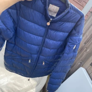 Blå Moncler dunjacka med dragkedja - Snygg blå dunjacka från Moncler med klassisk quiltad design. Jackan har dragkedja framtill, två fickor med dragkedja och en extra ficka på ärmen. Perfekt för kyliga dagar och ger en clean look med sin smala passform och blanka finish. Lite slitage och litet litet hål vid armen längst ner vid handleden, och har inte ärlighetsbevis på den pga att jag fick den i present och därav varför den säljs billigt!