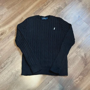 Svart kabelstickad tröja Polo Ralph Lauren - Kabelstickad svart tröja från Polo Ralph Lauren med klassisk rund halsringning och vit broderad logga på bröstet. Tröjan har lång ärm och ribbade muddar för en snygg passform. Perfekt för dig som gillar stilren och tidlös design.