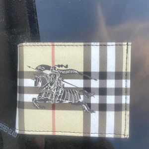 Rutig plånbok från Burberry - Snygg plånbok från Burberry med klassiskt rutigt mönster i beige, svart, vitt och rött. Framsidan har den ikoniska riddarloggan tryckt i svart. Plånboken är tillverkad i skinn och har en stilren, tidlös design som passar perfekt i fickan.