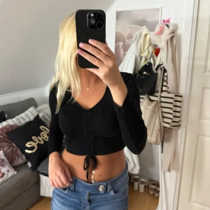 Svart croppad topp från ONLY - Svart långärmad croppad topp från ONLY med v-ringning och snörning framtill. Toppen är ribbad och sitter tight, perfekt till jeans eller kjol. Enkel men ändå trendig design som passar till många olika stilar.