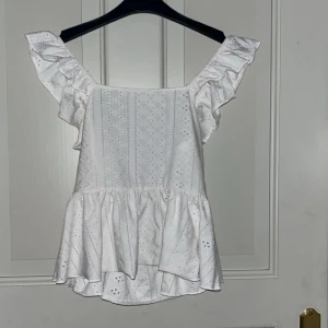 Vit volangblus med broderi och knyt - Supersöt vit blus med volangärmar och broderade detaljer. Toppen har fyrkantig ringning, volanger längs axelbanden och en luftig, flowy passform. Baksidan har snygg knytning i midjan för extra detalj. Perfekt för sommarens alla tillfällen!