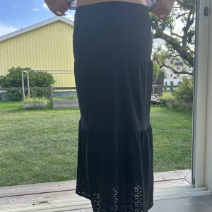 Svart långkjol från Kappahl - Svart långkjol från Kappahl med broderade håldetaljer och volang nertill. Kjolen har en mjuk och flowy känsla, perfekt för dig som gillar en chill och stilren look. Resår i midjan för extra komfort.