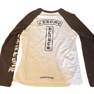 Chrome Hearts longsleeve - Säljer en långärmad tröja från Chrome Hearts med vita och mörkgrå kontrastärmar. Stora tryck med Chrome Hearts-logga på ryggen och ärmarna. Skön passform och tillverkad i bomull, perfekt för dig som gillar streetwear vibes.
