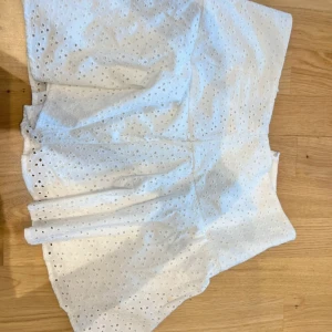 Vit kortkjol i broderad bomull - Supersöt vit kortkjol i bomull med broderade hål och lätt volang. Perfekt för varma dagar och ger en fräsch och somrig vibe. Kjolen har hög midja och är riktigt flowy och luftig.