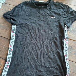 Svart t-shirt från FILA med logoband - Svart t-shirt från FILA med korta ärmar och rund hals. Snygga logoband med FILA-tryck i vitt, rött och blått längs sidorna. Tillverkad i mjuk bomull, perfekt för en sportig och avslappnad stil.
