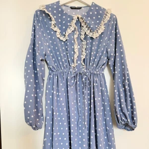 Blå prickig klänning med spetskrage - Supersöt blå klänning med vita prickar från Stein. Har stor spetsprydd krage, knytband i midjan och långa puffärmar. Klänningen har romantiska vibbar och är perfekt för dig som gillar vintageinspirerad stil.