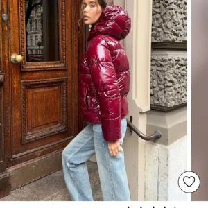 Vinröd glansig pufferjacka   - Säljer en vinröd pufferjacka med glansig finish och stor huva. Jackan har dragkedja framtill och breda, quiltade partier som ger en riktigt puffig look. Perfekt för kalla dagar.
