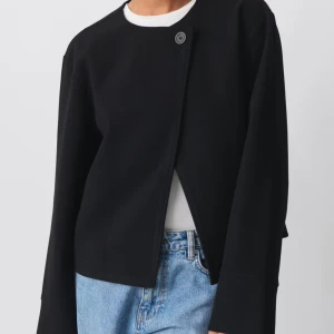 Svart kappa med asymmetrisk knäppning - Stilren svart kappa med rak passform och en unik asymmetrisk knäppning med en knapp vid halsen. Jackan har rund halsringning och långa ärmar, perfekt för en clean och modern look.