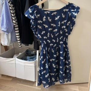 Blå kortklänning med fågelmönster - Supersöt blå kortklänning med vitt fågelmönster och smock i midjan. Klänningen har volangärmar och en luftig känsla, perfekt för varma dagar. Den har en avslappnad passform och är tillverkad i ett lätt material.