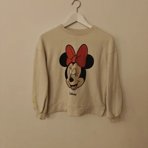 Disney sweatshirt - Beige sweatshirt från Disney med stort tryck av Minnie Mouse på ryggen. Motivet har röda och svarta detaljer med glittriga paljetter på rosetten och ansiktet. Tröjan har rund hals, långa ärmar och ribbade muddar. Perfekt för dig som älskar Disney och vill sticka ut.