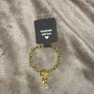 Guldigt armband med pärlhänge - Snyggt armband i guldtonad metall med chunky länkar och ett hänge i form av en rosett och en droppformad pärla. Perfekt accessoar för att lyfta din outfit med en söt och trendig detalj.