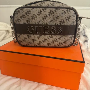 Beige axelväska från Guess med logomönster - Snygg beige axelväska från Guess med tydligt logomönster och bred svart detalj med stor GUESS-text framtill. Väskan har silverfärgad kedjerem och dragkedja med Guess-logga. Perfekt storlek för det viktigaste och stilren insida med Guess-tryck.