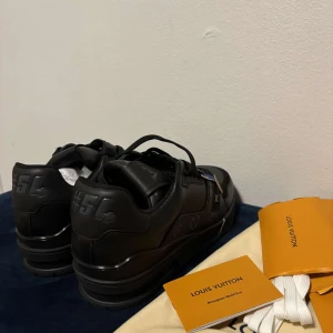 Louis Vuitton svarta sneakers monogram - Exklusiva svarta Louis Vuitton sneakers med monogramdetaljer och #54 på hälen. Skorna har rund tå, platt sula och är tillverkade i skinn med LV-logga på tungan. Snygga svarta skosnören och tydlig Vuitton-text på sulan. Kommer med dustbags och extra vita skosnören.