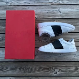 Valentino Open sneakers vit/svart  - Snygga Valentino Open sneakers i vitt skinn med bred svart läderdetalj över mitten. Klassisk rund tå, platt sula och diskreta nitar baktill. Kommer med original röd box. Perfekta för dig som gillar stilrena och lyxiga sneakers.