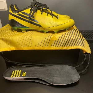 Gula adidas fotbollsskor med svarta detaljer - Snygga gula adidas fotbollsskor med svarta ränder och svarta snören. Du får med kvitto,extra sulor och påsen.