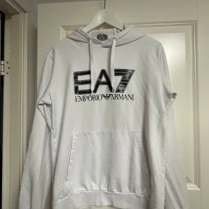 Vit hoodie från Emporio Armani EA7 - Snygg vit hoodie från Emporio Armani EA7 med stor logga framtill. Klassisk känguruficka, huva med snörning och långa ärmar. Perfekt för en clean och stilren look. Mjuk bomullskänsla och relaxed passform.