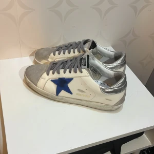 Golden Goose Superstar sneakers med blå stjärna - Golden Goose Superstar sneakers i vitt skinn med grå mocka på tån och grå snörning. Sidorna har en blå stjärna och hälen är metallic-silver. Platt svart sula och GGDB-logga på plösen. Snygg kontrast mellan färger och material, perfekt för dig som gillar streetstyle. Box och dustbag kommer med. Skriv om ni har några frågor!