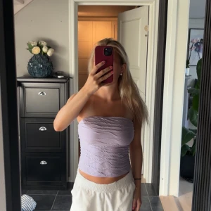 Ljuslila bandeau tubtopp - Säljer en ljuslila bandeau tubtopp med snyggt rynkad struktur. Toppen är axelbandslös och har en croppad passform som sitter tight mot kroppen. Perfekt för varma dagar och enkel att styla med dina favoritjeans eller kjol.