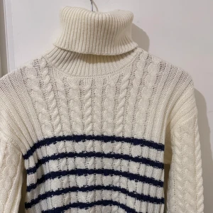 Vit polotröja med blå ränder - Säljer en chunky stickad polotröja i vit med mörkblå horisontella ränder. Tröjan har grova kabelstickade detaljer och en hög, ribbad polokrage. Långärmad och perfekt för kyliga dagar. Snygg att styla med jeans eller kjol för en chill look.