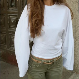Vit långärmad topp med vida ärmar - Säljer en vit långärmad topp med riktigt breda och vida ärmar. Modellen är slim och sitter snyggt runt midjan, vilket ger en clean och modern look. Materialet känns som mjuk bomull och toppen är helt utan mönster eller detaljer. Perfekt för dig som gillar minimalistisk stil.