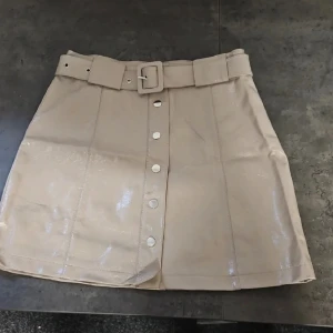 Beige kortkjol med bälte och knappar - Snygg beige kortkjol med glansig finish och markerade sömmar. 