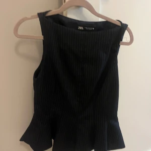 Svart randig linnetopp från Zara - Snygg svart ärmlös linnetopp från Zara med tunna vita ränder och volang nertill. Toppen har en rak halsringning och figurnära passform som ger en cool och dressad look. Perfekt att styla med jeans eller kjol för en trendig outfit. Finns ej att köpa på Zara längre.