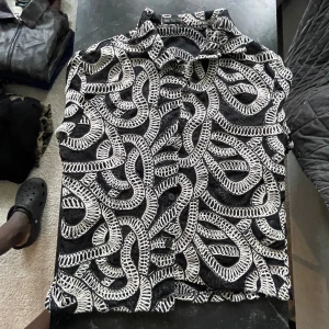 Svart skjorta med vit broderad design - Unik svart skjorta med vit broderad mönster som slingrar sig över hela plagget. Skjortan har klassisk krage och knappar framtill. Materialet känns som bomull och broderiet ger en cool och konstnärlig vibe. Perfekt för dig som gillar att sticka ut med din stil.