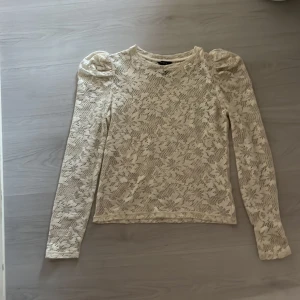 Vit spetsblus med puffärm från Lindex - Säljer en vit spetsblus från Lindex i storlek S. Blusen har lång ärm med snygga puffar vid axlarna och är helt täckt av ett blommigt spetsmönster. Perfekt för dig som gillar romantiska detaljer och vill ha en feminin look.