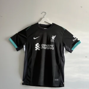 Liverpool FC svart Nike Diogo J. tröja - Liverpool FC fotbollströja med Diogo Jota och nummer 20 på ryggen. Svart med vita tryck och turkosa detaljer på ärmarna. Nike Dri-Fit material som andas, officiell Standard Chartered sponsorlogga och LFC-emblem på bröstet. Perfekt för dig som vill ha en snygg supportertröja. Rip Jota