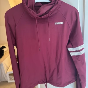 Vinröd träningströja från Stronger - Säljer en vinröd träningströja från Stronger med vita ränder på ena ärmen och logga på bröstet. Tröjan har snörning både vid halsen och nederkanten, samt långärmad design. Materialet är mjuk bomull med lite stretch.