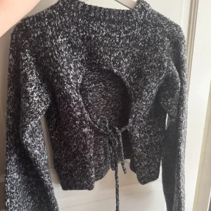 Grå stickad tröja med öppen rygg NA-KD - Säljer en mörkgrå och vitmelerad stickad tröja från NA-KD i storlek S. Tröjan har lång ärm, croppad passform och en cool öppen rygg med snörning. Endast använd någon enstaka gång!