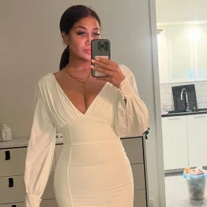 Vit långklänning med slits och v-ringning - Säljer en elegant vit långklänning med djup v-ringning och långa puffärmar med knappar vid ärmslutet. Klänningen har en figurnära passform och en hög slits på ena sidan som ger en snygg touch. Perfekt för dig som vill sticka ut med stil.