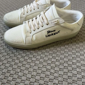 Saint Laurent vita sneakers strl 42 - Säljer ett par vita Saint Laurent sneakers i canvas med handskriven logga på sidan. Skorna har rund tå, platt sula och snörning. Snygg minimalistisk design med diskreta slitningar och detaljer i skinn. Perfekt för dig som gillar exklusiva streetwear-vibbar.