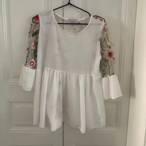 Vit blus med broderade blommor - Superfin vit blus från Lovelio med transparenta ärmar och rygg, dekorerade med broderade blommor i rött, rosa, grönt och lila. Blusen har en rund halsringning, volang vid midjan och trekvartsärmar med breda manschetter. Perfekt för dig som gillar romantiska detaljer!